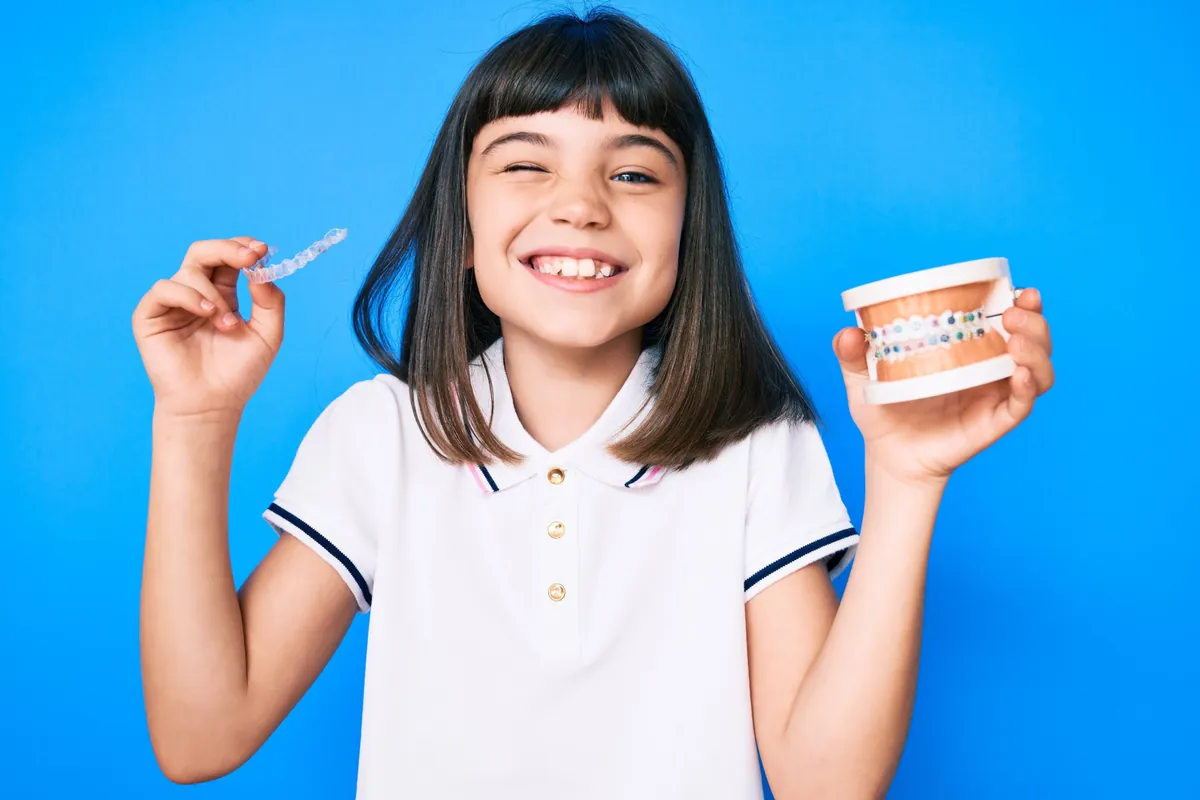 Smiling child holding a clear aligner and a braces model